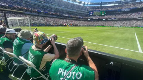 Simon Stacpoole A row of people in green photographer bibs, each pointing cameras at a football pitch inside a very large stadium
