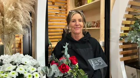 Florist Vicky Hudspith-Bailey at her stall. She is smiling at the camera and has flowers displayed in front of her.