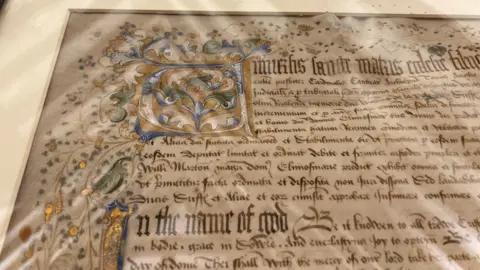 The lefthand corner of the original Almshouse charter is shown. The text is Medieval English and written with flourishes. It is ornately inscribed in gold and blue and green. You can make out "In the name of God" at the start of the second paragraph.