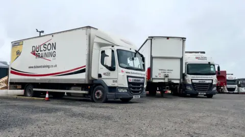 Three white lorries parked on a gravel yard. "Dulson Training" is printed on the side, front and back of the vehicles.