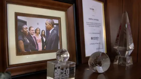 A desk with awards and a picture of Peju with President Obama