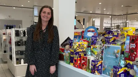 BBC Alana Rimeur standing to the left of a table top which has a range of Easter eggs on it. She is wearing a black and white polka dot dress and is smiling at the camera. In the background is the store with white shelving with items on them.