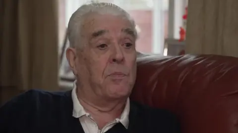 A man with short grey hair and a dark coloured jumper and white top is sitting on a burgundy sofa. 