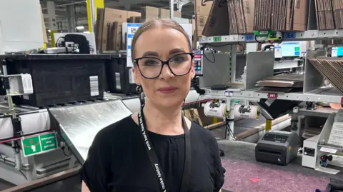 Kate Bradbrook/BBC Magda Ziavka who transferred from Marston gate, she is wearing a black shirt and lanyard. She is wearing large eye glasses with a black frame and in a warehouse surrounded by collapsed cardboard boxes which will be used for packagaing Amazon orders.