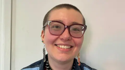 A woman with short hair wearing glasses and smiling at the camera. She has dangly earrings in her ears 