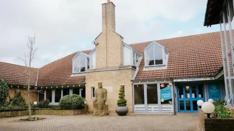 The hospice's building in Gosforth is brown brick with sculpted bushes at the front leading on to a court yard. There is a brown statue of a seated man. 
