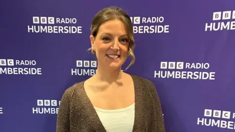 Grace Hunt. She has dark hair and is wearing a white top and brown cardigan. She is standing in front of a purple banner with BBC Radio Humberside logos 