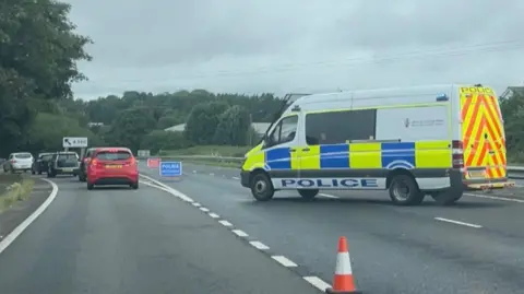 A police van is parked across a dual carriageway and cars are queuing as they leave the road via a slip road. There is a sign reading: POLICE ACCIDENT and a traffic cone is in the foreground. It is a grey day.