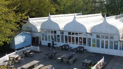 LDRS A white cafe building with four separate, pointed roof features that give way to windows and doors running along the ground floor. A paved area crowded with wooden benches sits outside the building. 