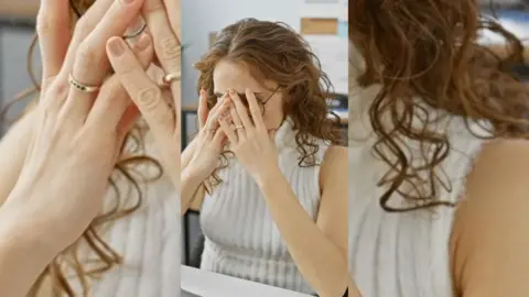 Woman with long brown hair, sat at a desk, covering her face with her hands.