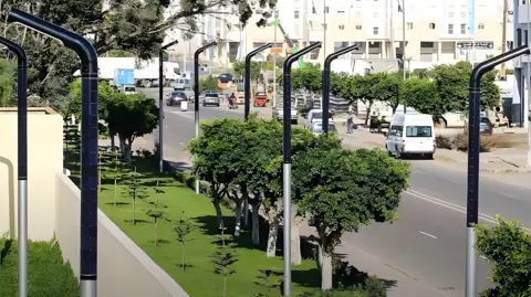 Conflow Power Group A picture of a tree-lined street in Morocco with iLamps visible in green spaces next to the road an on a terrace. The lamps are curved like a bendy drinking straw and the top half is wrapped in solar cells. 