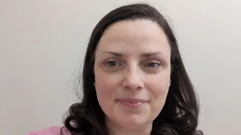 Maria smiles as she poses for selfie against a white wall. She has dark hair and wears a pink top.