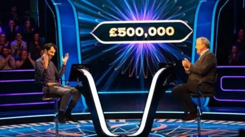 ITV Two men sitting down opposite each other. The man on the left is wearing brown trousers and a brown shirt and has his hands in the air. The man on the right is wearing a brown suit and clapping. There is a sign with £500,000 in white lettering in the background. There are people sat down on either side clapping.