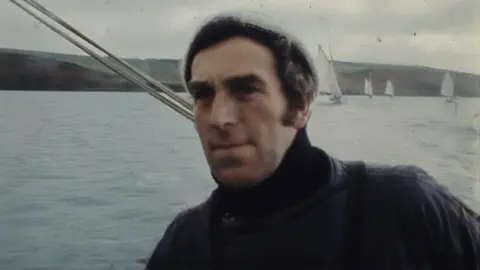 Paul Mulraney A grainy photo that shows Paul Laity on a boat looking past the camera. He is at sea and there are working boats under sail and a headland behind him. He has dark hair and is  wearing a woolly hat and a sou'wester. The sea is calm and the sky is grey with cloud.