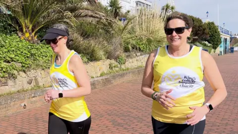 Two women in yellow tops with 'King's College Hospital Charity' on them run along the Eastbourne seafront.