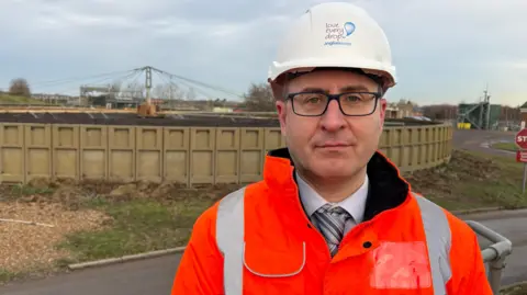 Ben Schofield/BBC Geoff Darch, standing outside in a hard hat and orange high-visibility jacket and looking directly down the camera. He is wearing a tie and shirt underneath the high-vis jacket. He is standing towards the right of frame and behind him is a 