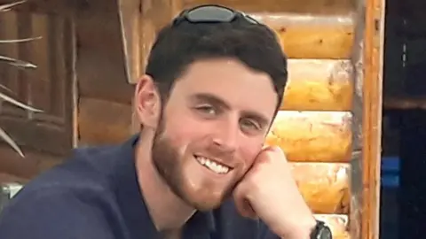 Andrew Harper smiles, wearing a dark blue shirt and resting his race on his hand