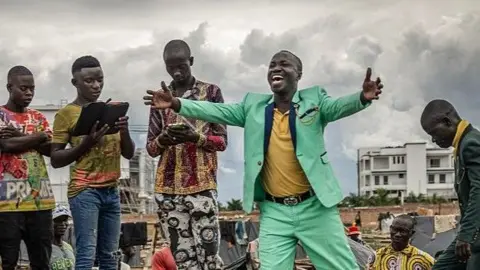A man in a green suit on top of a boat, preaching to a crowd.