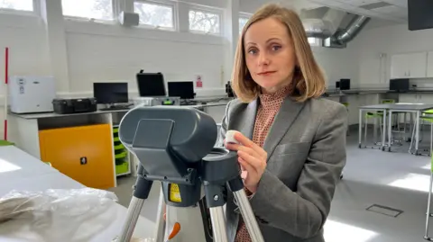 A woman in a grey jacket and a red shirt looks at the camera as she stands over a piece of technical metal equipment with four legs