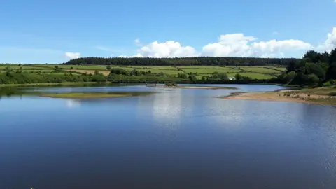 The reservoir has an island in the middle surrounded by green banks, with fields and trees in the distance. The photograph was taken on a sunny day, with blue skies.