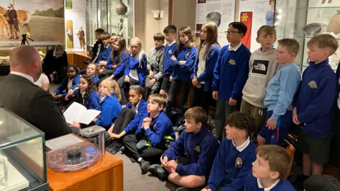 Vikki Irwin/BBC A large group of school students wearing blue jumpers standing in a museum. They are sitting down and standing up. They are looking at a man who is sat in front of them reading a book.
