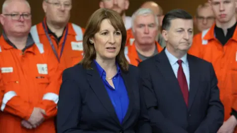 PA Media The Chancellor Rachel Reeves and Scottish Secretary Douglas Alexander at the Grangemouth plant. Ms Reeves has short brown hair and a blue jacket and top. Mr Alexander is wearing a blue suit and maroon tie. Workers from the factory in orange clothing stand behind them