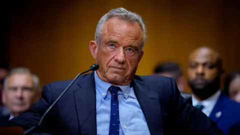 A Getty Images RFK Jr. fotografou em sua audiência no Senado. Ele está vestindo uma gravata azul real fina e uma camisa de botão azul claro com uma jaqueta de suíte azul de marinho escuro GA. Ela se bronzeou, com cabelos curtos. O público por trás disso está borrado. 