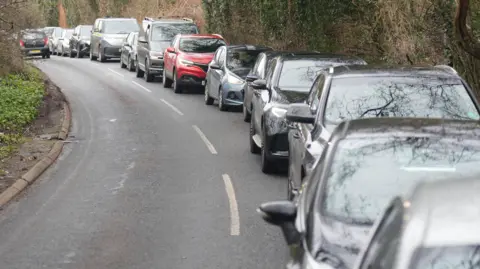 Eddie Mitchell Vehicled queueing up behind one another on a road facing towards the camera. A vehicle drives away from the camera on the other side of the road, which is clear.