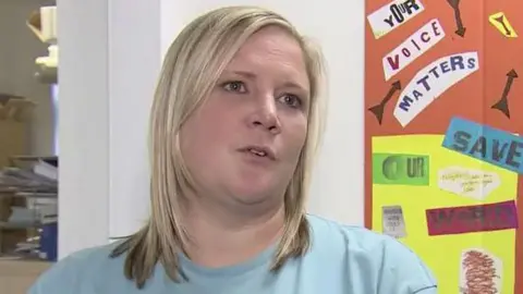 Head and shoulder shot of a young woman with shoulder length blonde hair. She is wearing a light blue sweatshirt emblazoned with a DAY logo. Behind her is a noticeboard with brightly coloured posters stating slogans including "your voice matters", "people before profits and "save our world".