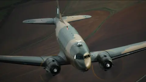 Darren Harbar/Crown Copyright BBMF Dakota plane shot from above with its nose towards the bottom of the image. It is painted in blue and cream camouflage colours. Beneath it are brown fields. 