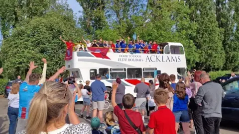 A white open-top double decker bus with football players in blue waving from the top. Several people can be seen waving in front of it and clapping.