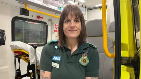 A paramedic in a green paramedics uniform is sitting in an ambulance smiling. She has long dark hair and tattoos.