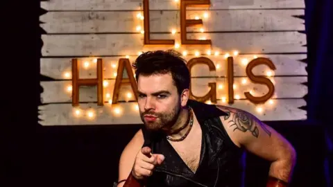 Big Burns Supper A man with dark hair and a beard and a rose tattoed on his shoulder points at the camera with a sign saying Le Haggis lit up behind him
