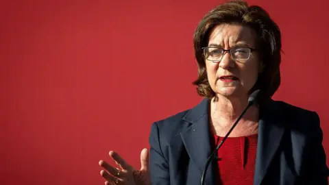 Matthew Horwood/Getty Images Eluned Morgan gesturing towards the camera, stood in front of a microphone. She is wearing glasses and is in front of a red background