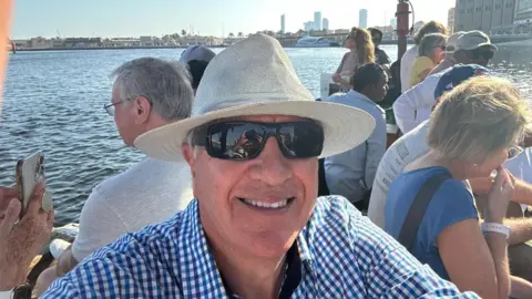 Terry O'Dell A selfie of Terry O'Dell who is sitting on a boat full of people. He is wearing a cream trilby hat, dark glasses and a blue and white gingham check shirt.