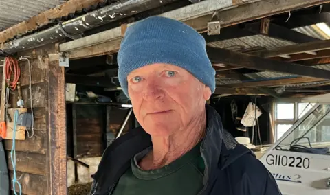 Steve Wale is stood in the middle. He is wearing a blue had and a navy coat and is stood in a wooden building. 