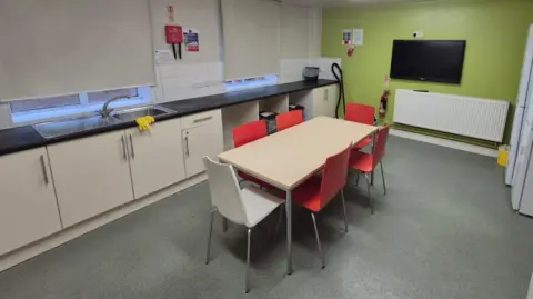 West Northamptonshire Council A kitchen with a table, four red chairs, a white chair and a green wall with a television on it. There is a white radiator and kitchen cupboards. There are fridges on the right. The floor is a greenish-grey. Blinds are pulled down at the two windows.