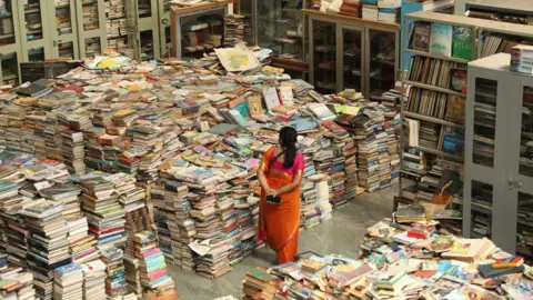Alphonse Vimulraj A woman walks through a room covered with piles of books. There are also numerous books stacked on shelves and in cabinets.