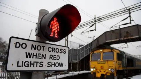 Getty Images Signs at a level crossing, that shows a red person on a traffic light above a sign saying "do not cross when red lights show". A freight train crosses underneath a footbridge nearby.