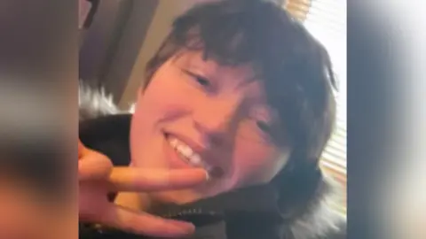 BBC Close up picture of Demi smiling looking at the camera. She has short brown hair and wears a black coat with a fur hood. She is doing a peace pose with her one hand. 