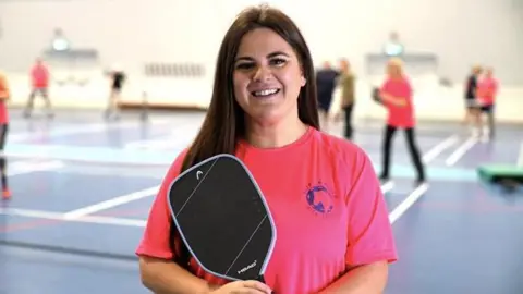 Jamie Niblock/BBC Christina is looking directly at the camera and is wearing the Suffolk Punch Pickleball T-shirt, which is bright pink with a blue logo. She is holding her pickleball paddle. There are other players in the background.