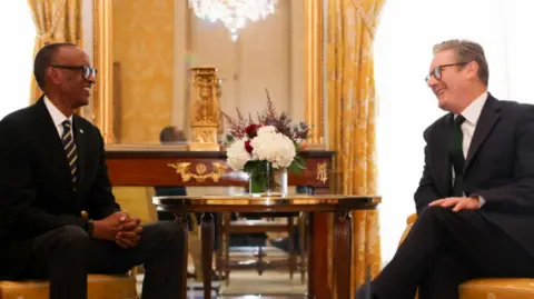 Rwanda President Paul Kagame and Prime Minister Keir Starmer pictured together in 2024. Both men are wearing suits and are sitting in a lavishly decorated room adorned with gold-coloured curtains and a table with flowers on it