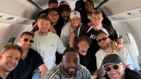 A group of football players are posing for a photo inside what looks like a private jet aircraft. Some are seated, others standing and they are smiling for the camera. Some are wearing sunglasses and have there arms around one another.