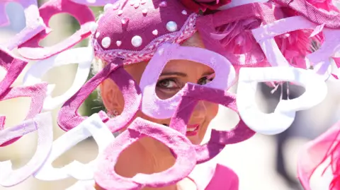 PA Media A woman looks through a gap in her headwear, which is a large fascinator with heart motifs coloured pink and white. She is smiling.