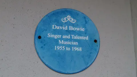 Heritage of London Trust A blue circular wall plaque mounted on a pale exterior wall with white text reading “David Bowie, Singer and Talented Musician, 1955 to 1968.”