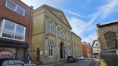 We can the 19th century church on a road in Norwich. Across the street we can see another church - and three cars are parked on the road. Above, is a bright blue sky with light cloud.