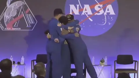 Artemis II astronauts hug on stage wearing blue overalls