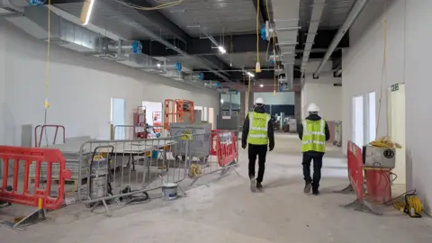Two men in yellow high-vis vest and hard hats walk through the Dome in Crewe. There is equipment around them.