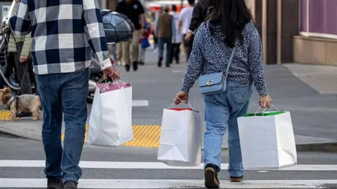 Shoppers cross a street holding white shopping bags.
