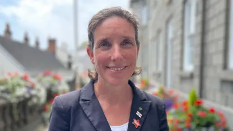 Deputy Lindsay de Sausmarez, a woman with brown hair wearing a blue jacket and three pun badges on the lapel. She is smiling towards the camera.